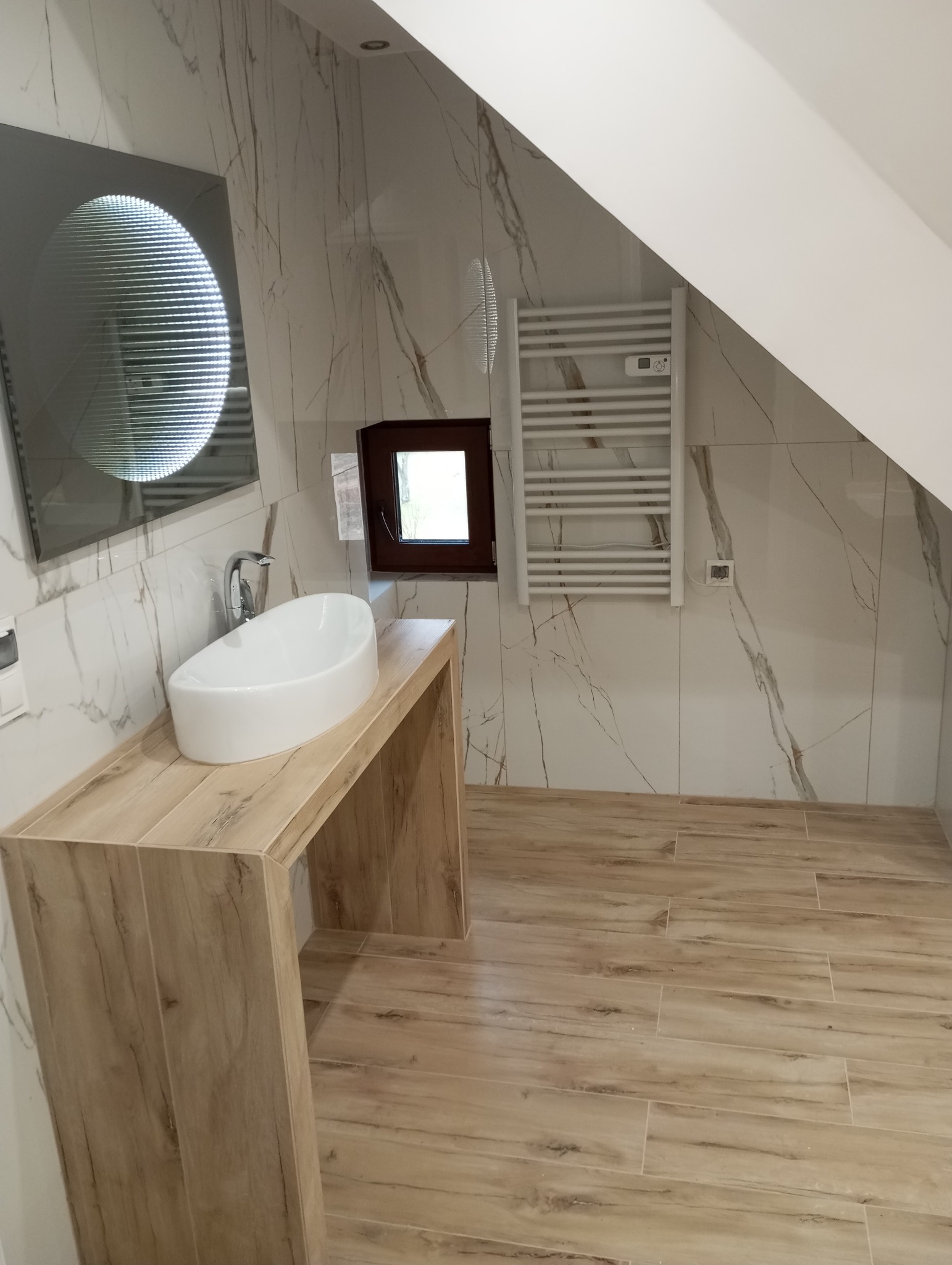 Nowoczesna łazienka na poddaszu z umywalką nablatową, grzejnikiem drabinkowym i podłogą imitującą drewno, oświetlona przez okrągłe lustro z efektem nieskończoności.