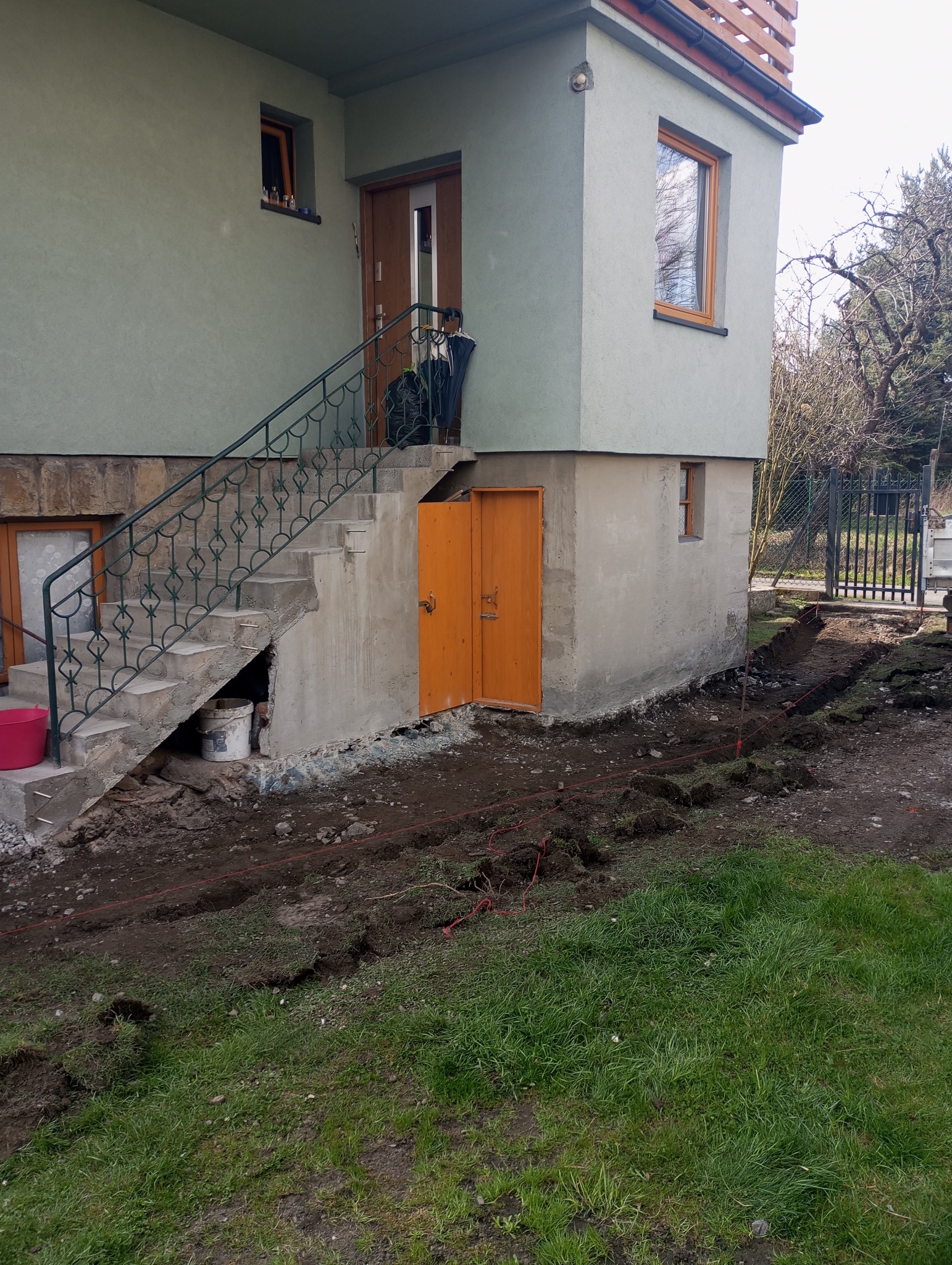 Przygotowanie terenu wokół domu z ocieploną elewacją, widoczne wykopane rowy pod izolację fundamentów, schody wejściowe z metalową balustradą, pomalowane na zielono ściany budynku.