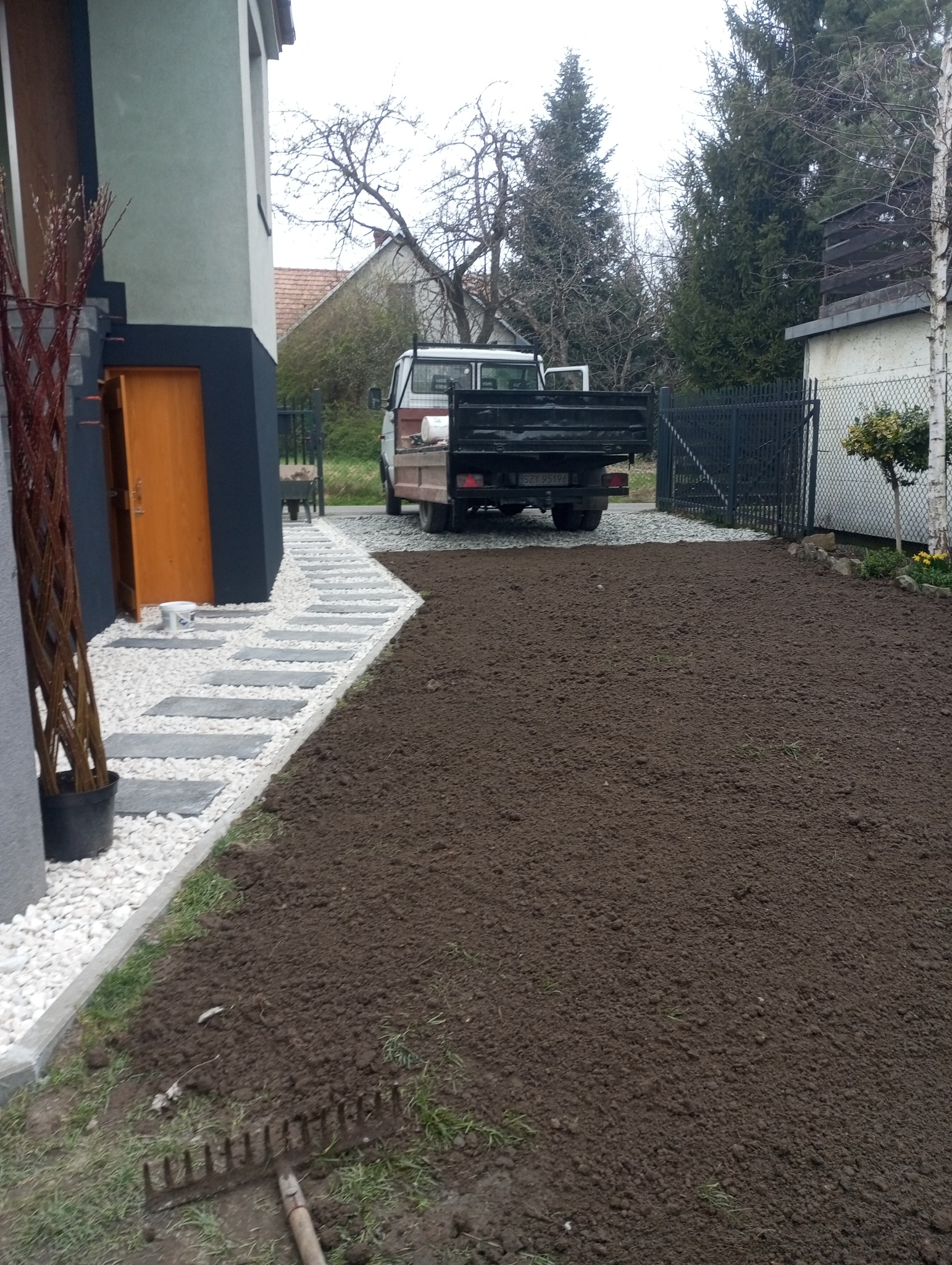 Przygotowany teren pod trawnik z widocznym grabiami, obok ścieżka z kostki brukowej i białego żwiru, w tle samochód dostawczy.