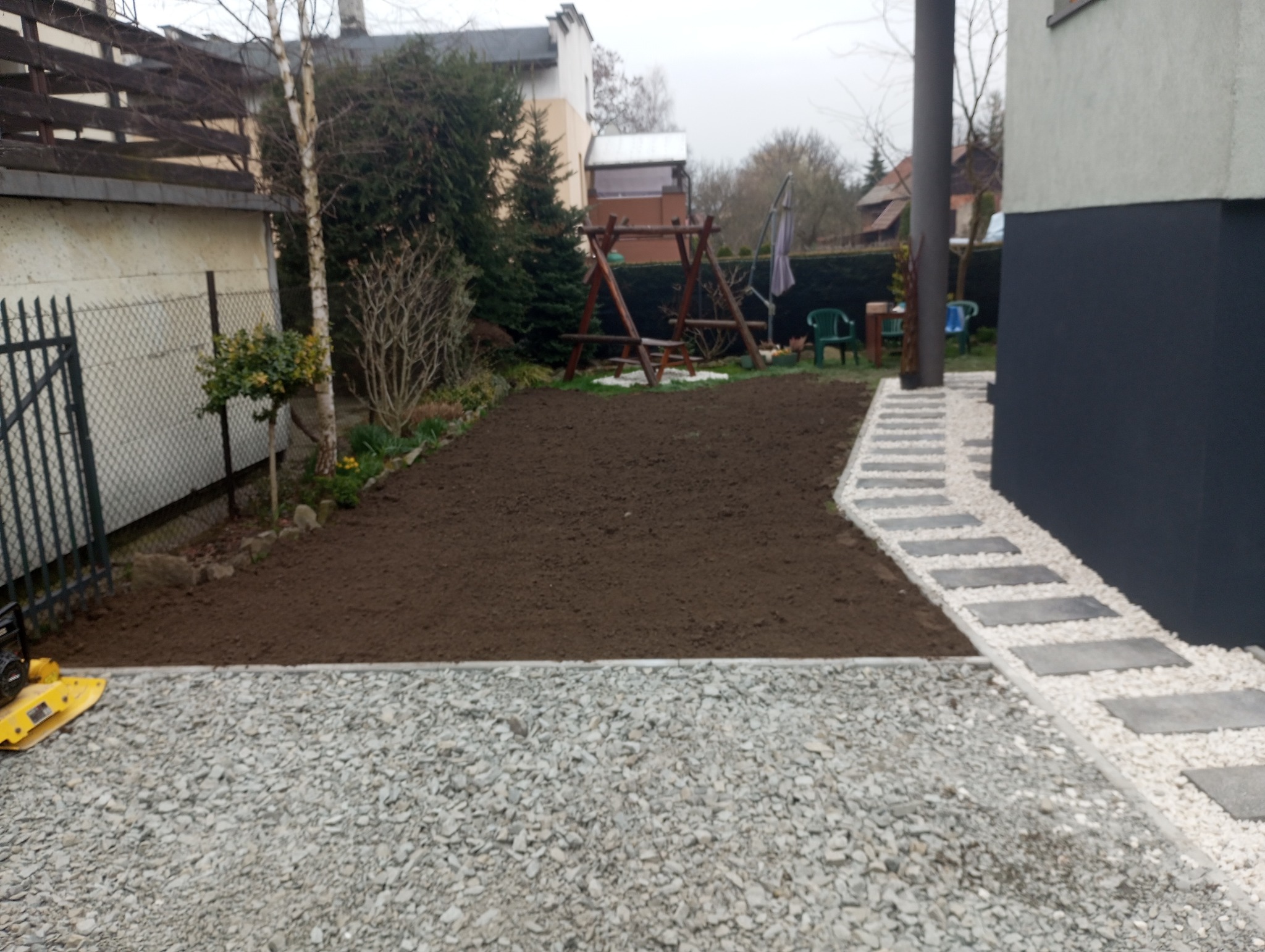 Przygotowany teren pod trawnik z nowo ułożonym chodnikiem z jasnych kamieni i płyt betonowych, widoczna zagęszczarka w tle, huśtawka ogrodowa.