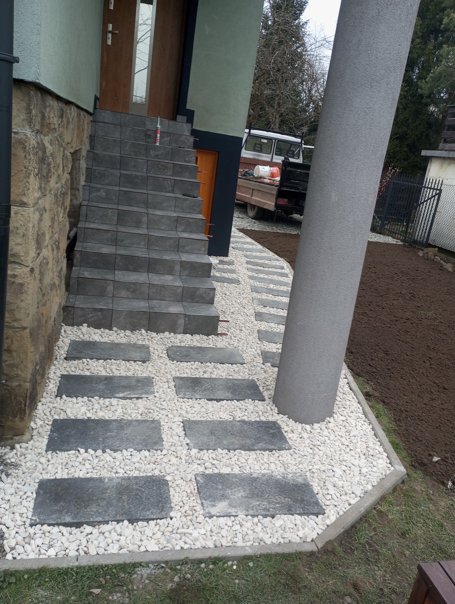 Ułożona ścieżka z szarych płyt na białym żwirze prowadząca do schodów wejściowych domu. W tle zaparkowany samochód dostawczy z narzędziami.