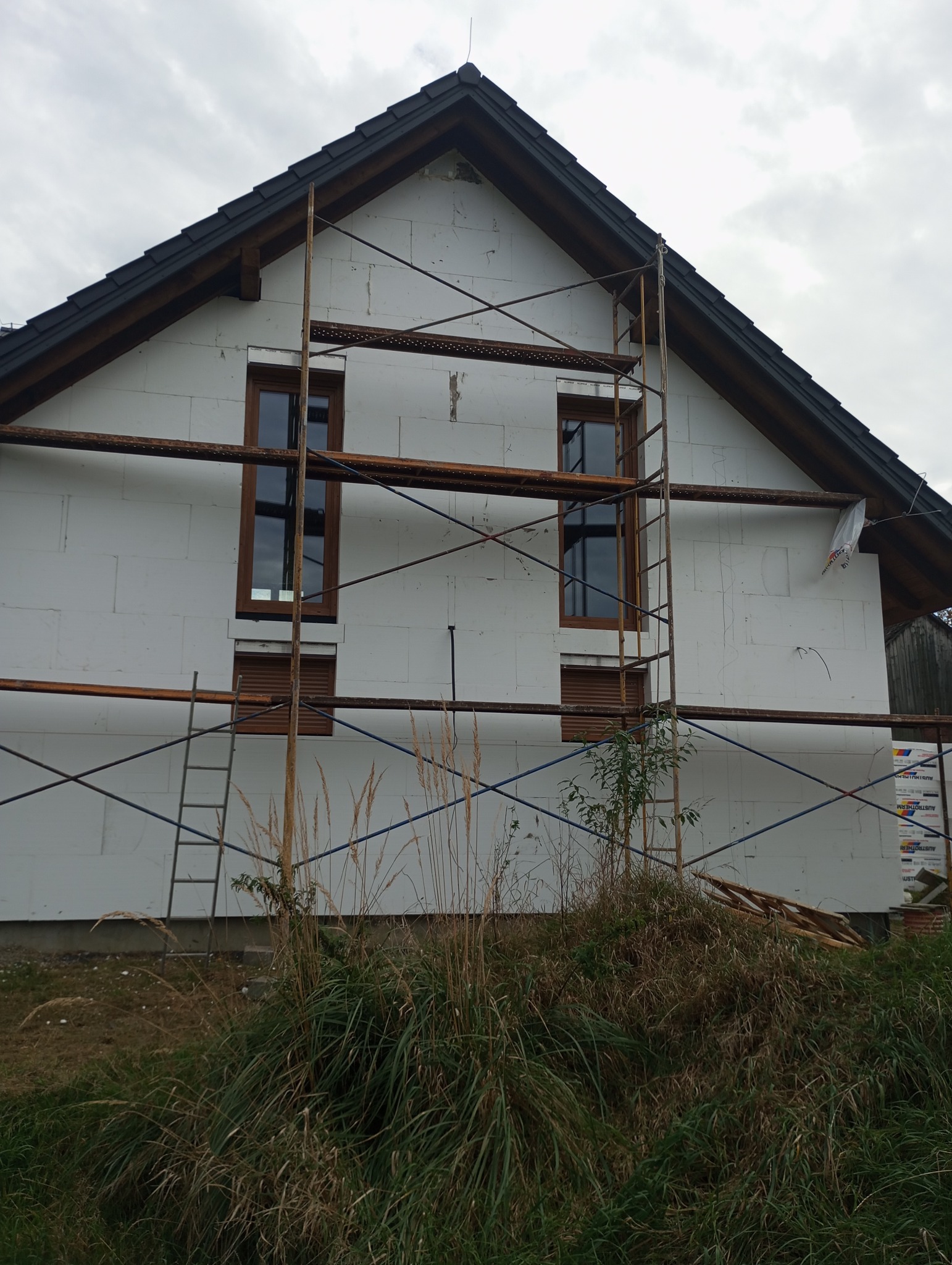 Dom jednorodzinny w trakcie ocieplania, widoczne rusztowania, styropianowe płyty izolacyjne na elewacji, nieukończone prace, zielona trawa i chwasty na pierwszym planie.