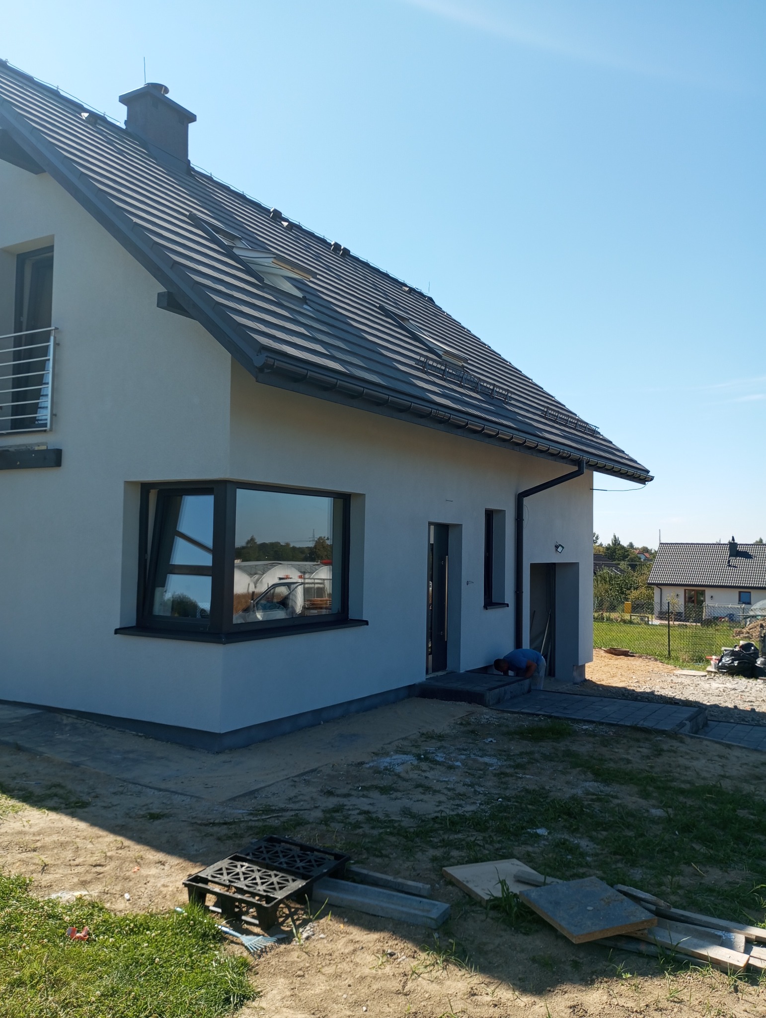 Nowoczesny dom jednorodzinny z szarą dachówką i oknem narożnym, elewacja w jasnym odcieniu, widoczny fragment układanej ścieżki z kostki brukowej, pracownik w trakcie układania