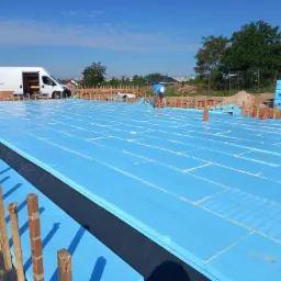 Izolacja fundamentu budynku w trakcie układania, widoczne niebieskie płyty styropianowe, drewniane szalunki, pracownik w oddali oraz dostawczy samochód w tle.