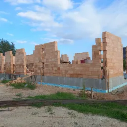 Wznoszenie ścian parterowego budynku z pustaków ceramicznych na ocieplonym fundamencie, widoczne zbrojenie i materiały budowlane.