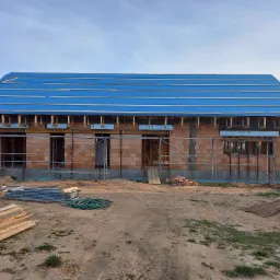 Niedokończony dom murowany z czerwonej cegły, z niebieską folią na dachu i rusztowaniami, widoczne materiały budowlane na placu budowy.