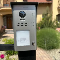 Szary wideo-domofon marki Vidos zamontowany na czarnym słupku ogrodzenia, z widoczną kamerą, głośnikiem, czytnikiem kart RF i przyciskiem dzwonka, na tle domu z beżową elewacją i czerwoną dachówką.