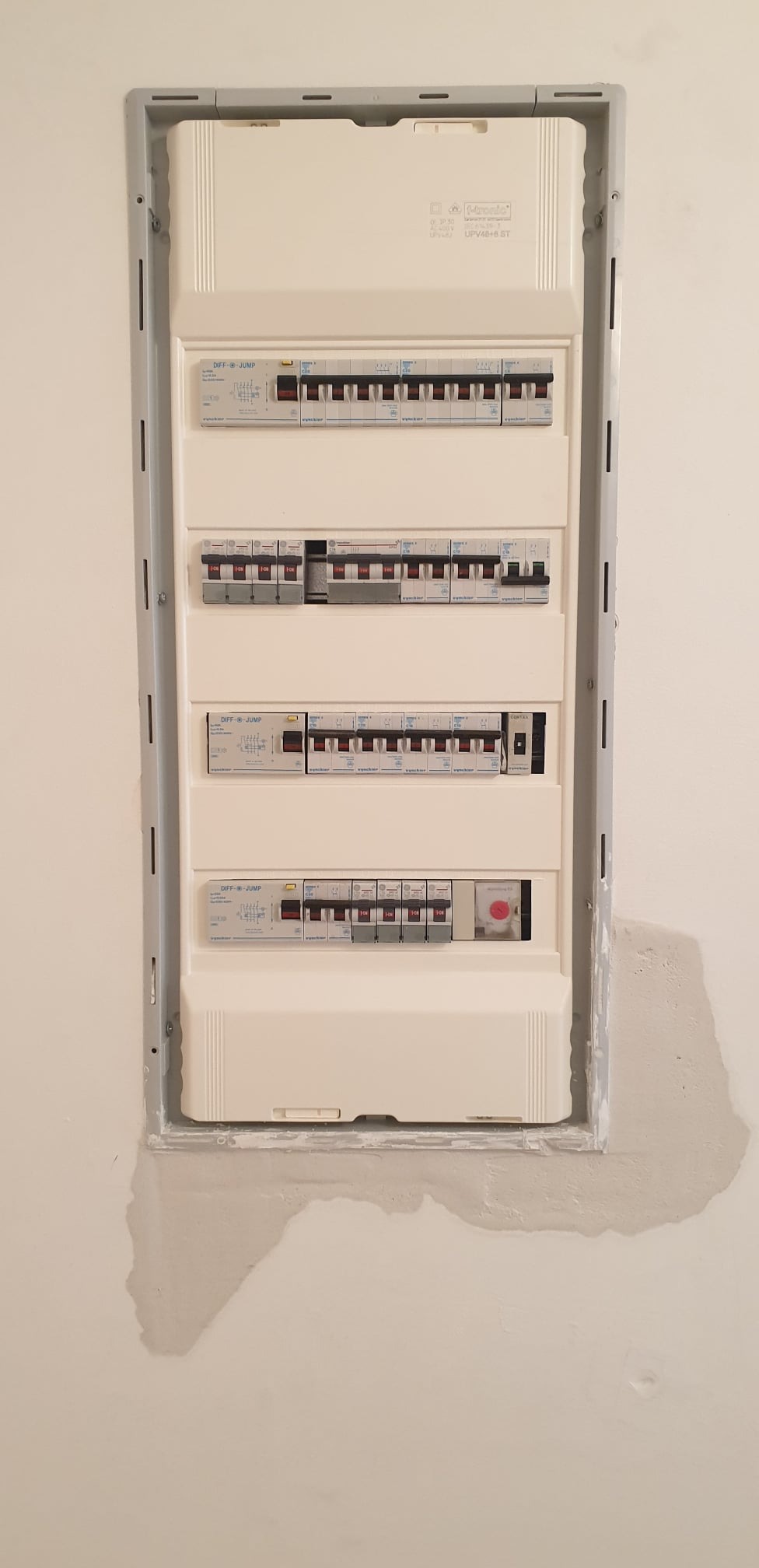 Rozdzielnica elektryczna z wyłącznikami różnicowoprądowymi i nadprądowymi, zamontowana w ścianie z widocznymi śladami po szpachlowaniu.