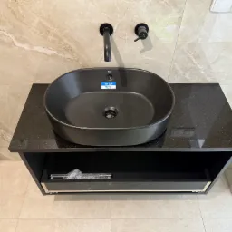 Czarna, owalna umywalka Roca na blacie z ciemnego kamienia z szufladą, zamontowana na ścianie z beżowymi płytkami imitującymi marmur. Minimalistyczna bateria.