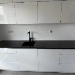 Biała, nowoczesna kuchnia z czarnym blatem kamiennym i minimalistyczną armaturą. Widoczne gniazdka elektryczne i odbicia w połyskujących frontach szafek.