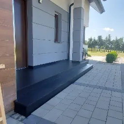 Nowoczesny taras wejściowy z ciemnymi schodami kamiennymi i szarą kostką brukową, widok na zadbany ogród z drzewami i krzewami, w rogu fragment drewnianej ramy okiennej.