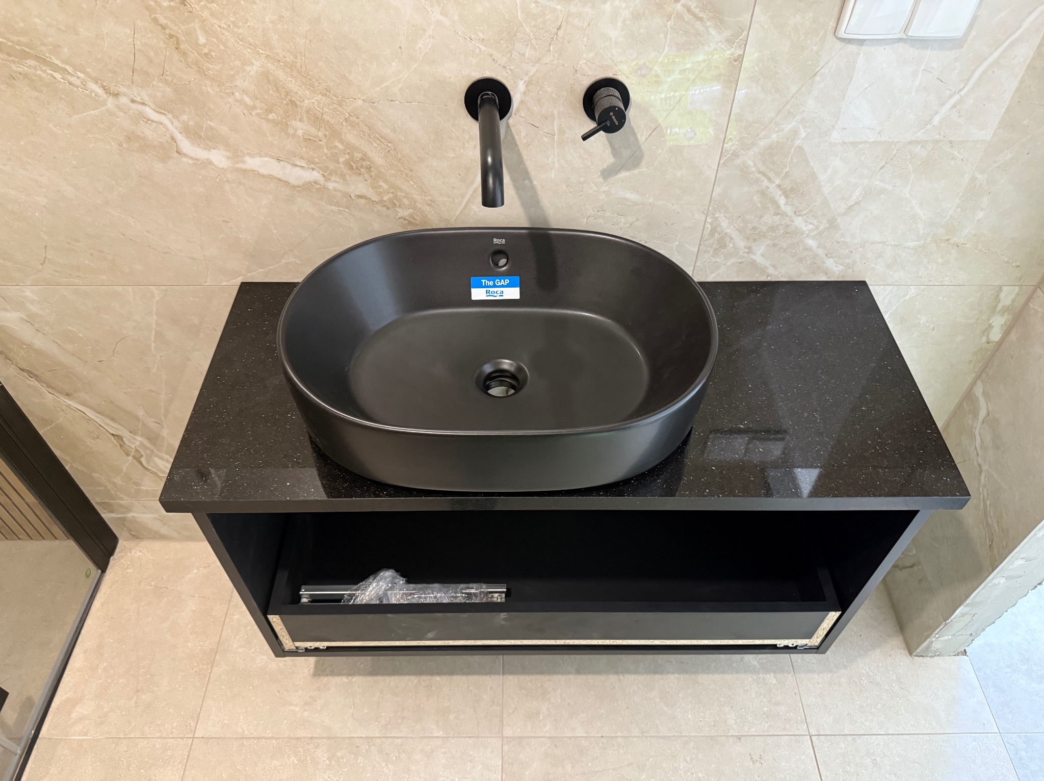 Czarna, owalna umywalka Roca na blacie z ciemnego kamienia z szufladą, zamontowana na ścianie z beżowymi płytkami imitującymi marmur. Minimalistyczna bateria.