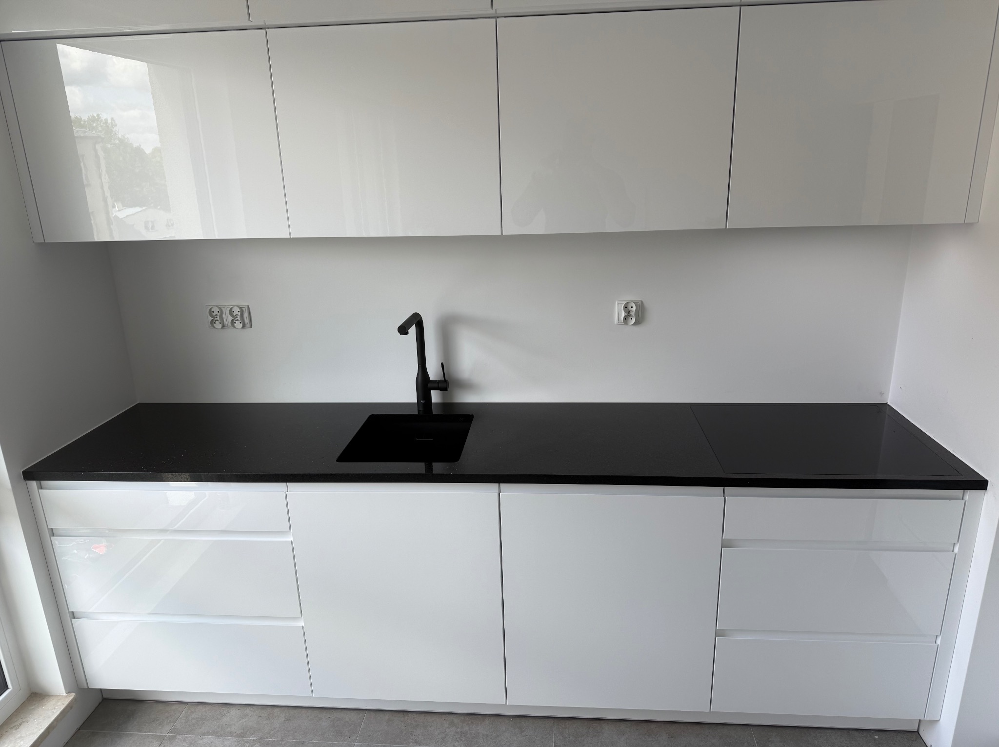 Biała, nowoczesna kuchnia z czarnym blatem kamiennym i minimalistyczną armaturą. Widoczne gniazdka elektryczne i odbicia w połyskujących frontach szafek.