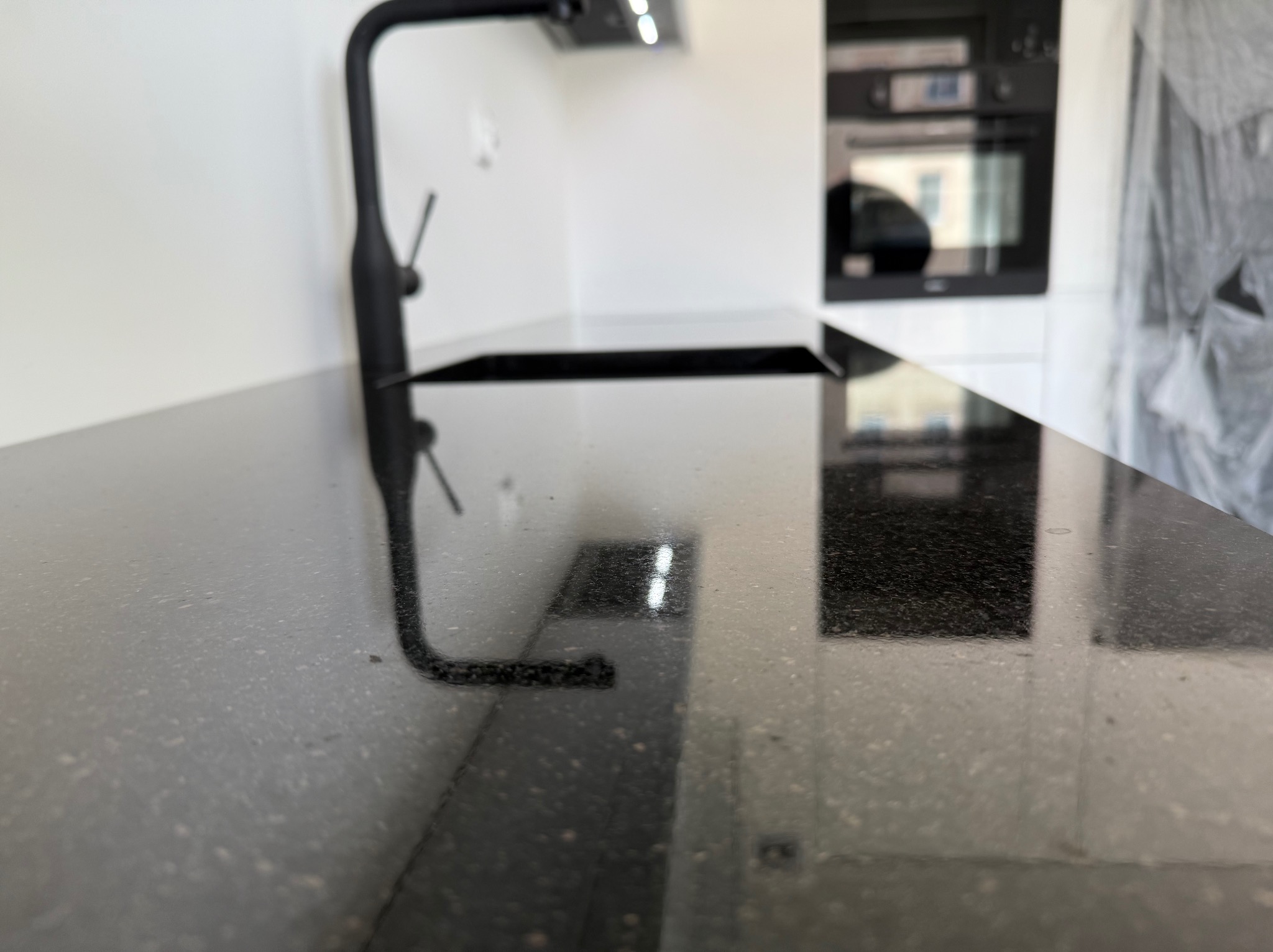 Czarny, błyszczący blat kuchenny z widoczną czarną baterią i płytą grzewczą, na tle białych szafek i piekarnika. Minimalistyczny design.