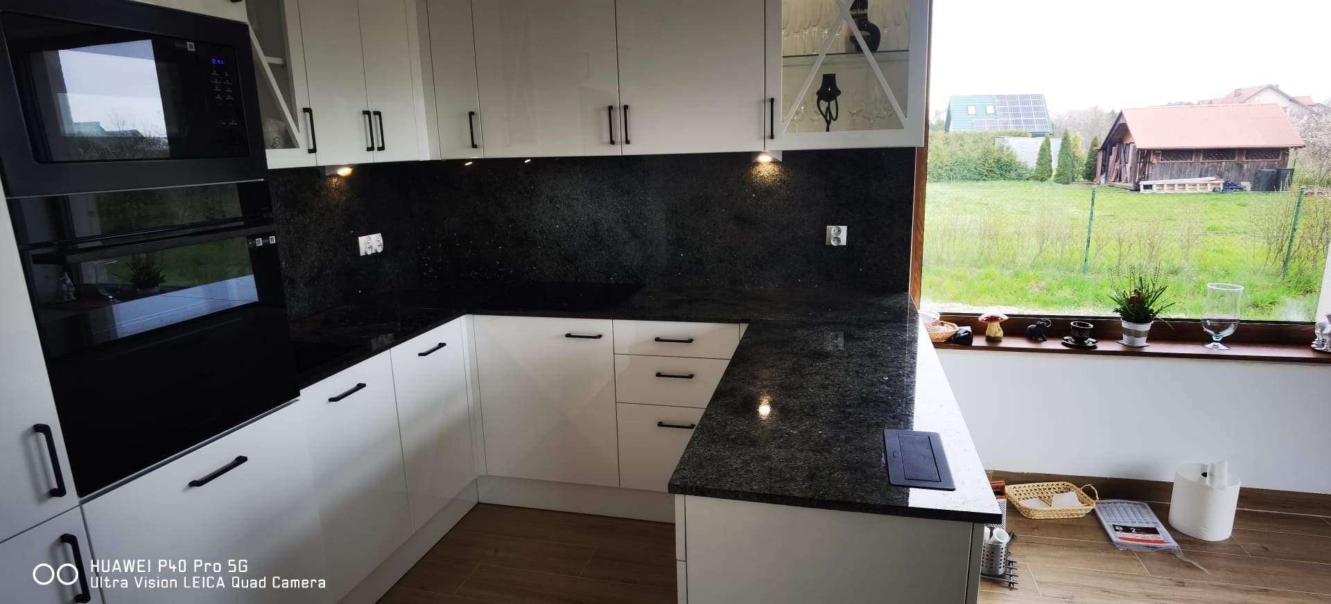 Błyszcząca, czarna powierzchnia robocza w nowoczesnej kuchni z białymi szafkami i czarnymi uchwytami, widok na ogród przez okno.