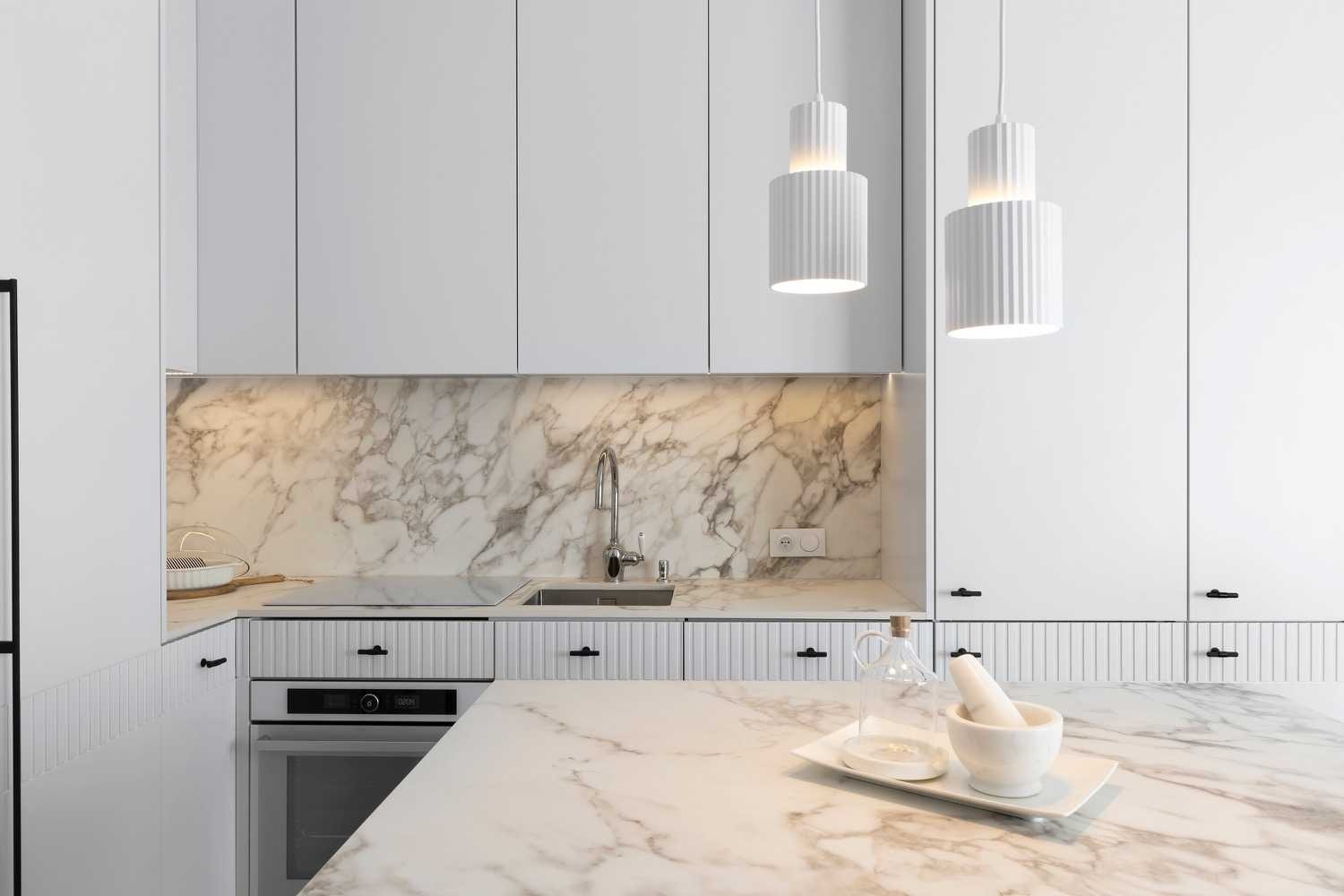 Biała kuchnia z marmurowym blatem, ścianą nad zlewem i wyspą, oświetlona wiszącymi lampami w kształcie walców z żłobieniami.