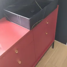 Czarna, prostokątna umywalka z marmurowym wzorem na czerwonej szafce łazienkowej z czterema szufladami i złotymi uchwytami, widoczna bateria umywalkowa.