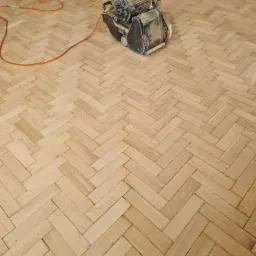 Parkiet ułożony w jodełkę, częściowo wyszlifowany maszyną do renowacji podł&oacute;g, widoczne ślady pyłu po obr&oacute;bce.