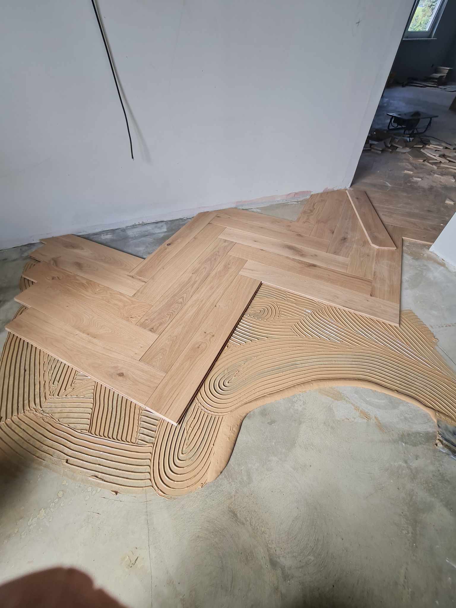 Układanie jasnego parkietu w jodełkę w Szczecinie, widoczny świeży klej na podłodze i fragmenty nieułożonych desek, w tle narzędzia i resztki materiałów.