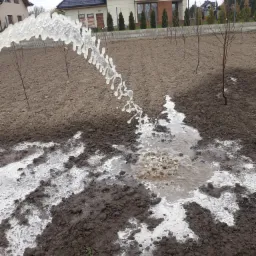 Woda tryskająca z ziemi w Warszawie podczas prac związanych z odwodnieniem terenu pod sad, w tle dom jednorodzinny.
