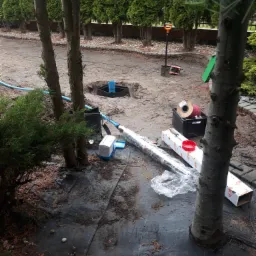 Teren przygotowany pod instalację, widoczny wykop z elementami studni, rury, skrzynka z kablem, narzędzia i czarna agrowłóknina rozłożona wokół, w tle rząd drzew iglastych.