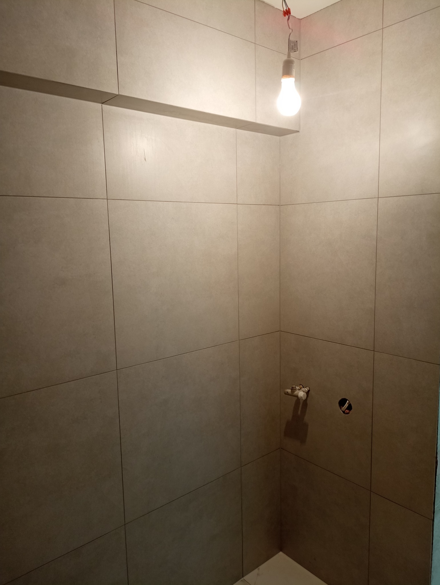 Świeżo położone, beżowe płytki ceramiczne w narożniku łazienki, z widoczną instalacją wodną i otworem na baterię, oświetlone wiszącą żarówką na kablu.
