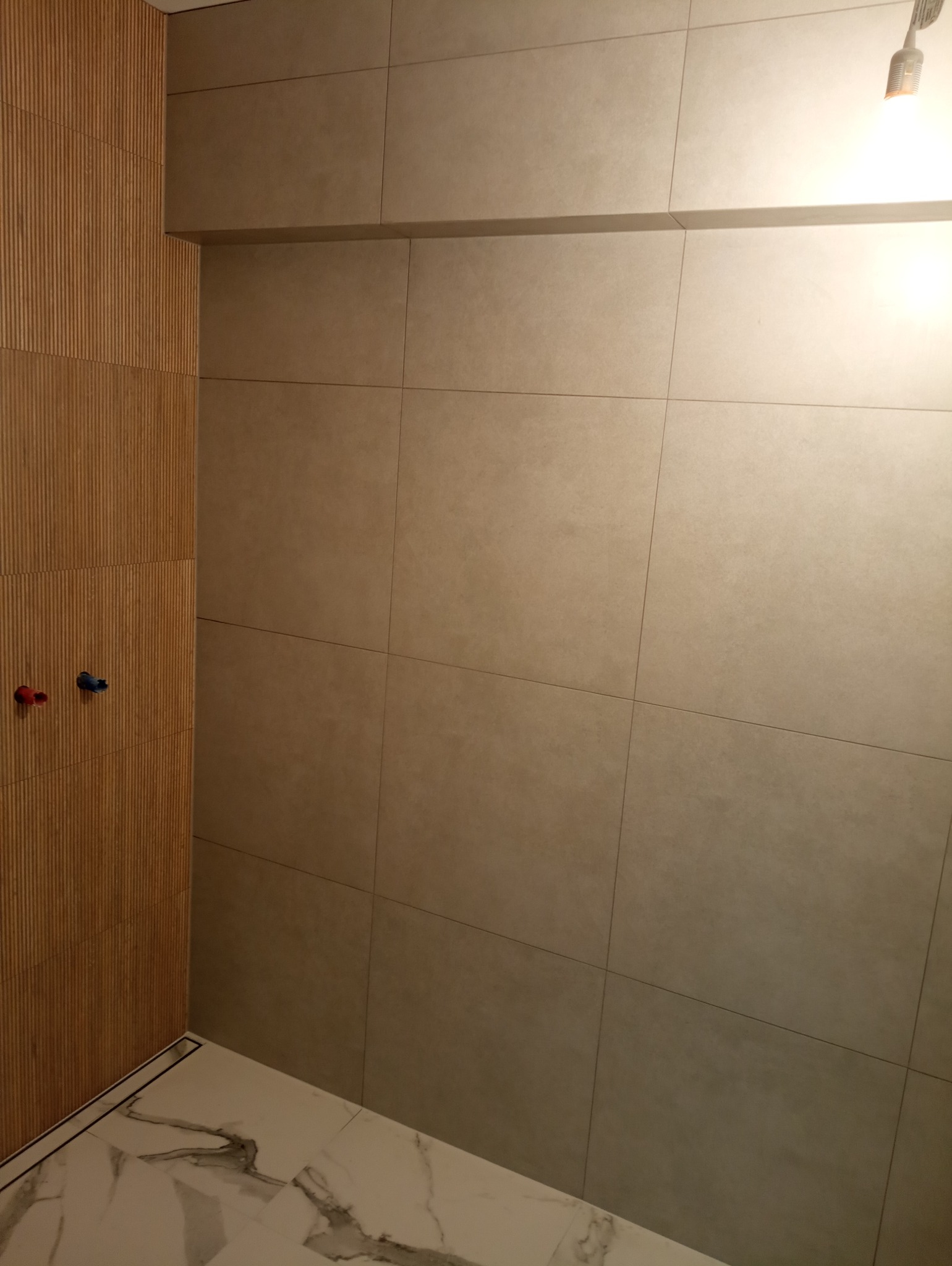 Wykończona łazienka z płytkami imitującymi drewno na jednej ścianie, szare płytki na pozostałych ścianach i marmurową podłogą z odpływem liniowym.