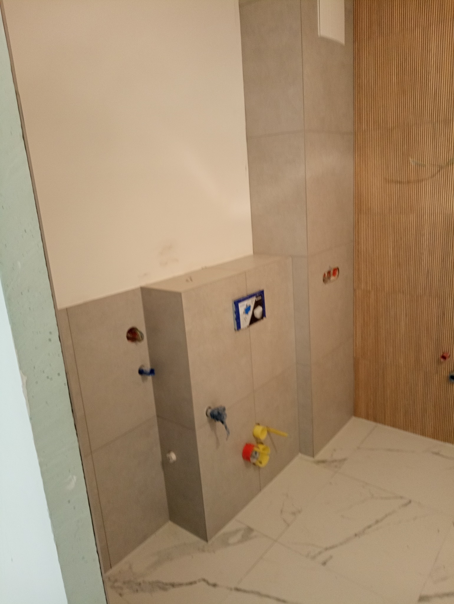 Wnętrze łazienki w trakcie remontu, widoczne płytki ceramiczne na ścianach, podłoga z marmurowym wzorem, przygotowane instalacje wodno-kanalizacyjne, drewniana boazeria na ścianie w tle.