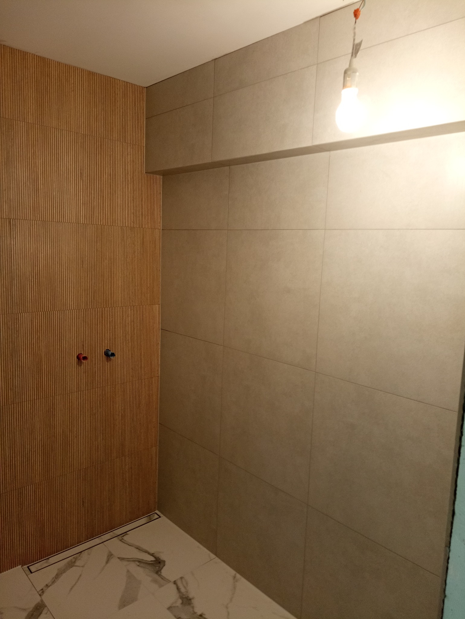 Wykończona łazienka z beżowymi, matowymi płytkami na ścianach, drewnianą okładziną z pionowymi lamelami, marmurową podłogą z odpływem liniowym i wiszącą żarówką.