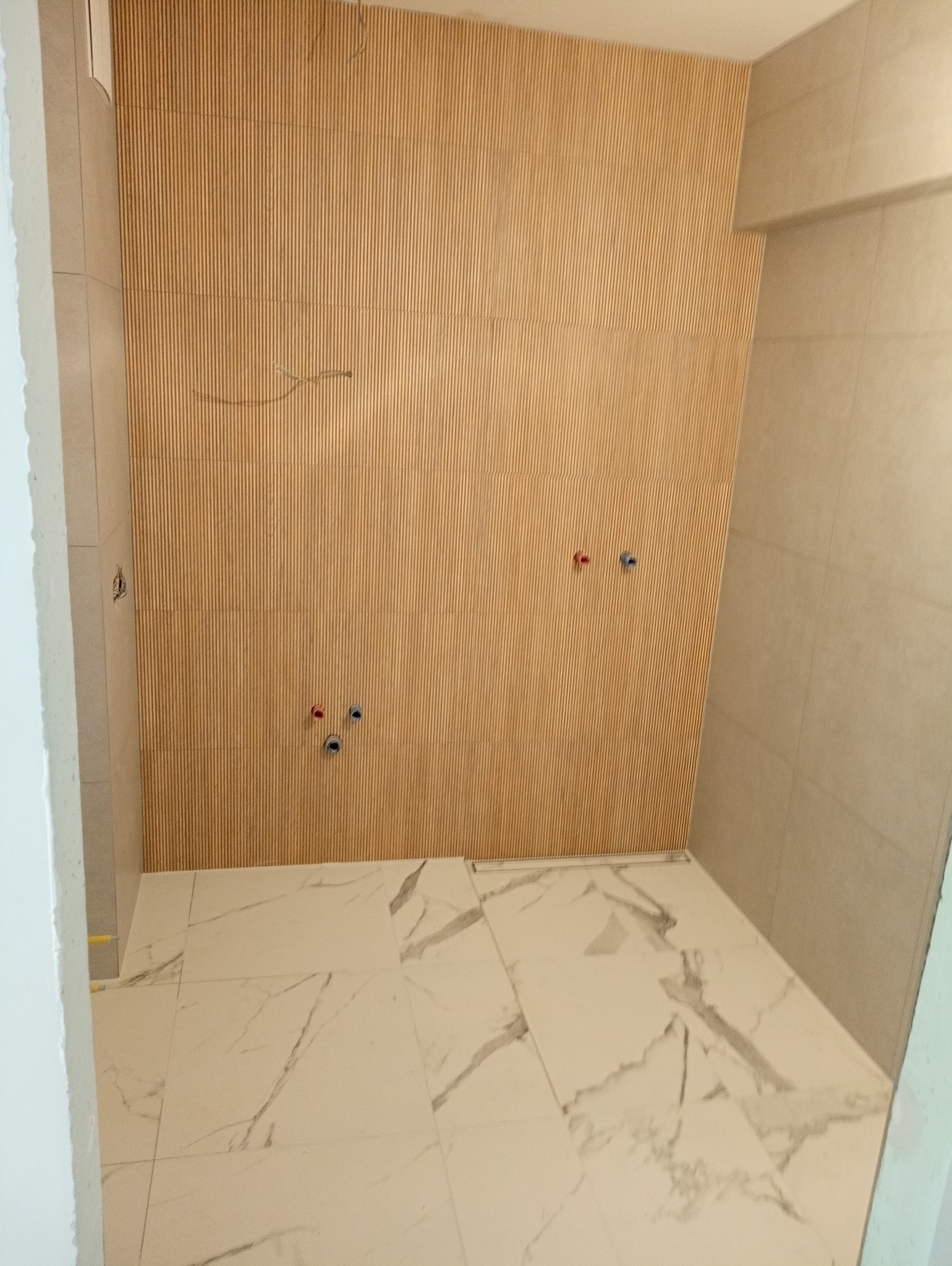 Łazienka w trakcie wykańczania: drewniane panele ścienne z widocznymi otworami na instalacje wodne, marmurowe płytki podłogowe z ciemnymi żyłami, jasne płytki na ścianach po prawej stronie.