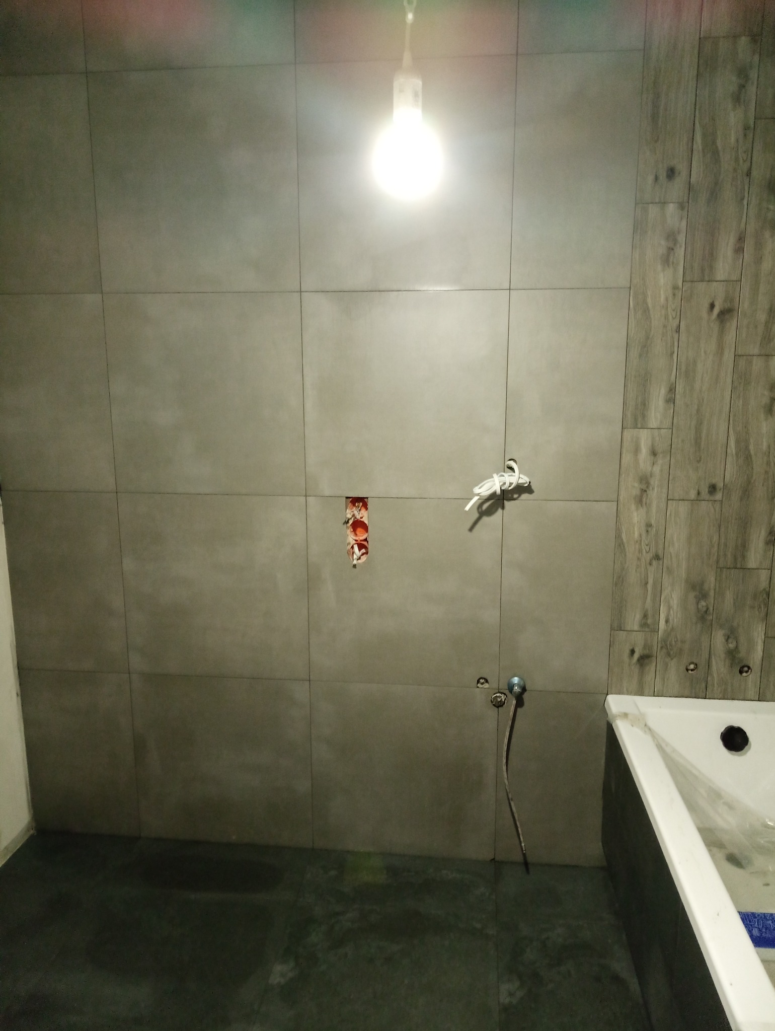 Łazienka w trakcie wykańczania: szare, kwadratowe płytki na ścianie, imitacja drewna na fragmencie ściany, widoczne przewody elektryczne i rura doprowadzająca wodę, surowa żarówka oświetlająca...