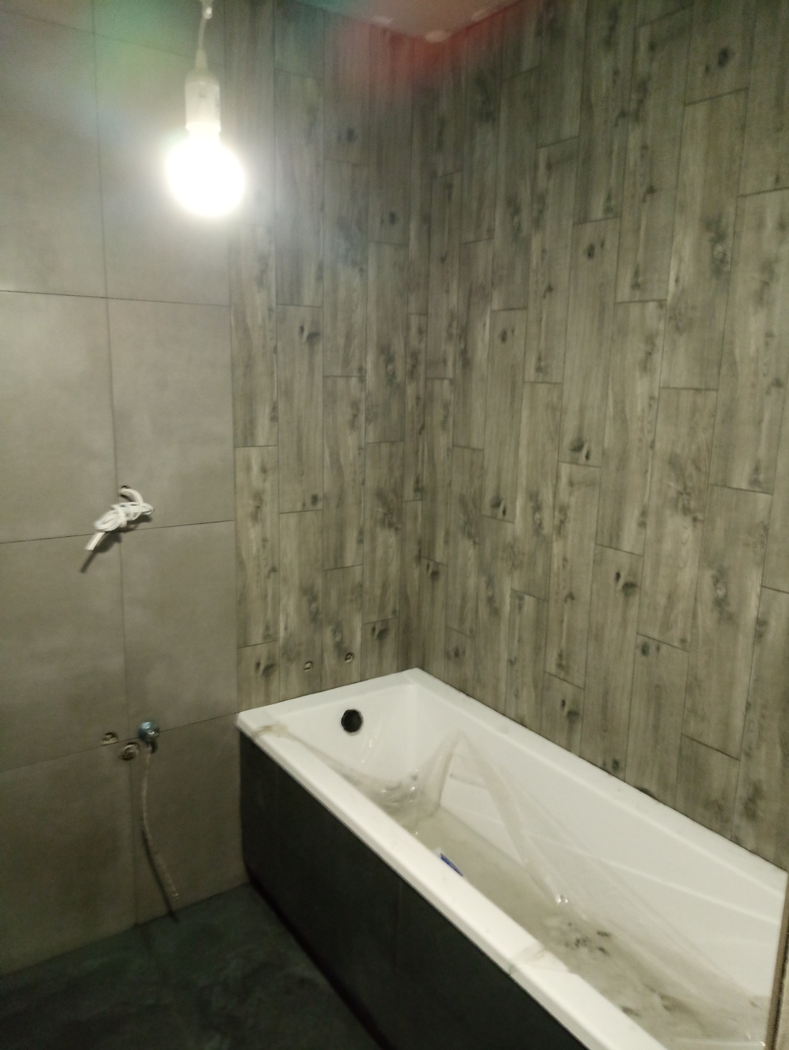 Łazienka w trakcie remontu: ściany wyłożone szarą, prostokątną glazurą imitującą drewno, biała wanna zabezpieczona folią, widoczne wystające kable i rury.