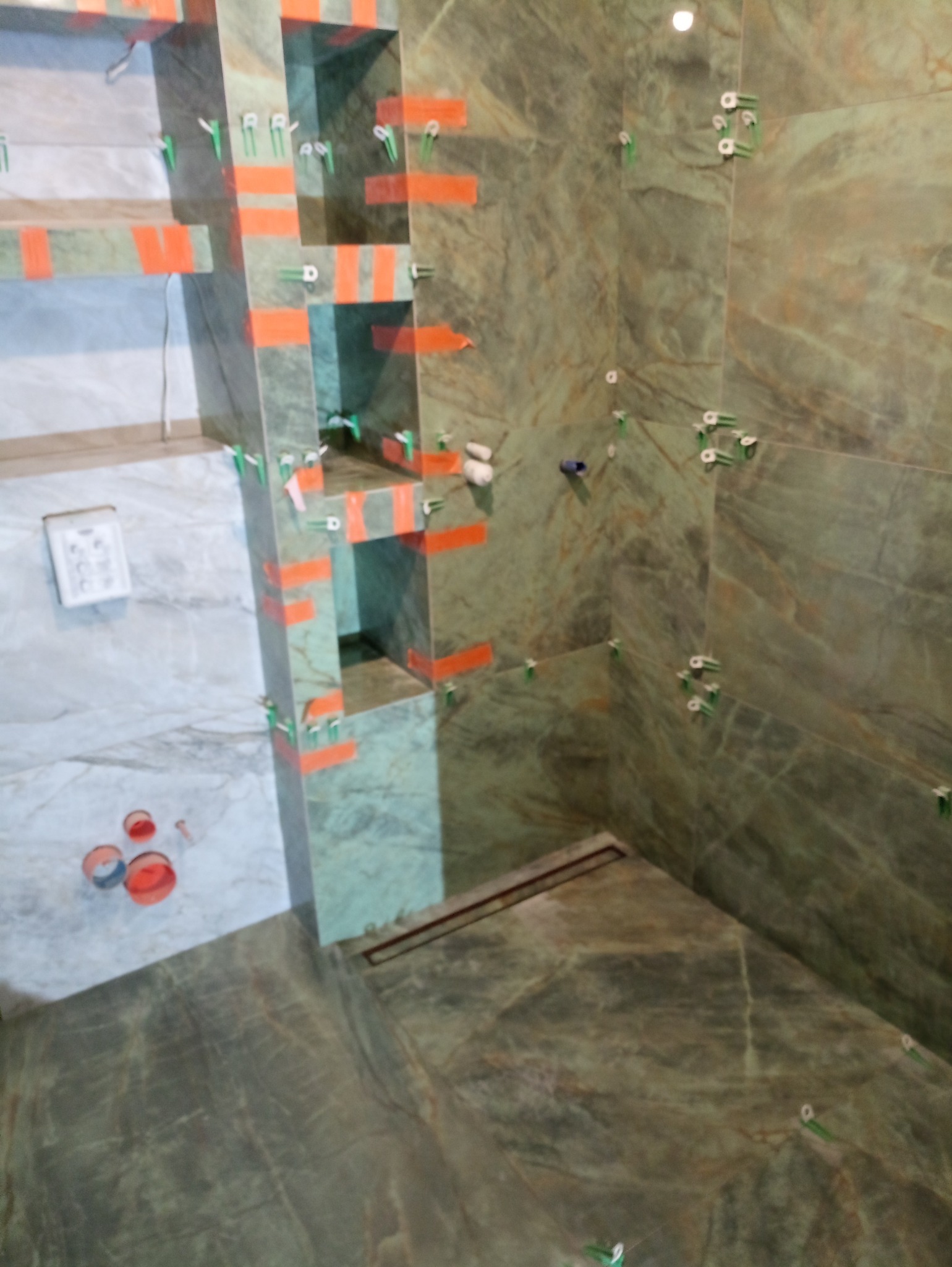 Montaż płytek imitujących marmur w łazience z wnękami, widoczne krzyżyki dystansowe i taśma malarska, odpływ liniowy w podłodze.