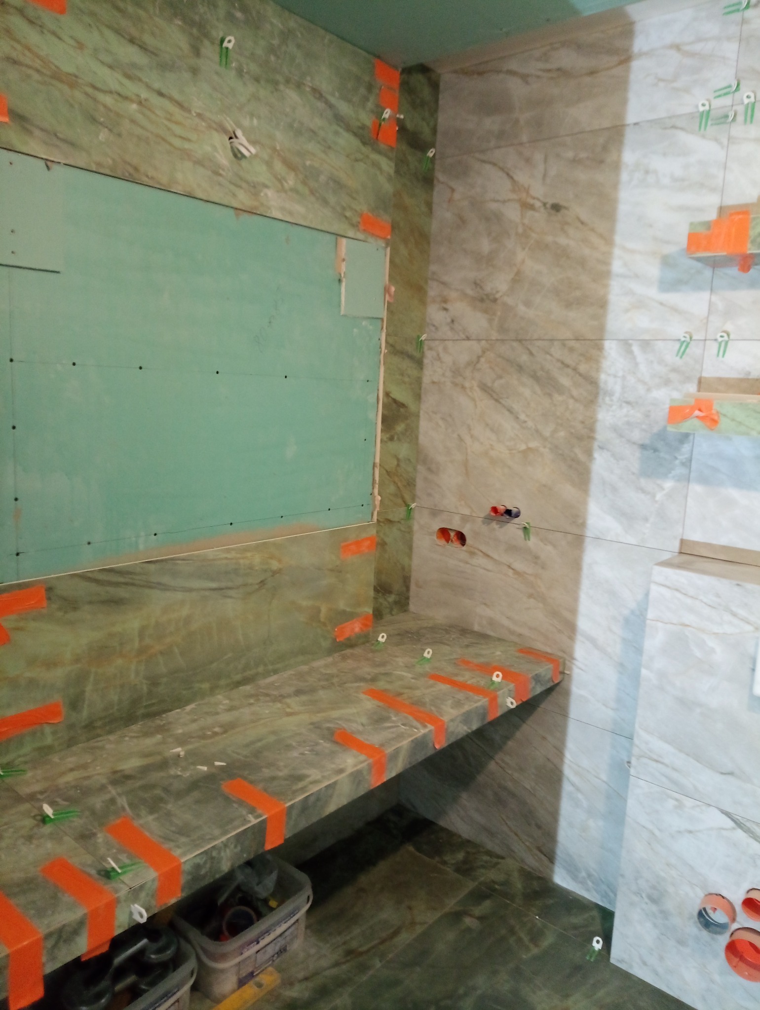 Montaż marmurowych płytek w łazience, widoczne płytki ścienne z marmurowym wzorem, fragment ściany z płytą kartonowo-gipsową, pomarańczowe kliny dystansowe i poziomica w wiadrze.