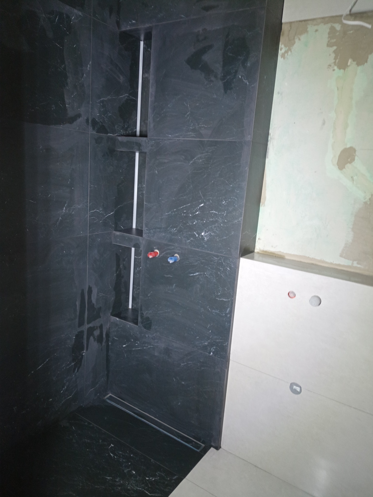 Czarne płytki imitujące marmur w kabinie prysznicowej z wnękami, widoczne podłączenia wodne, obok ściana w trakcie prac wykończeniowych.
