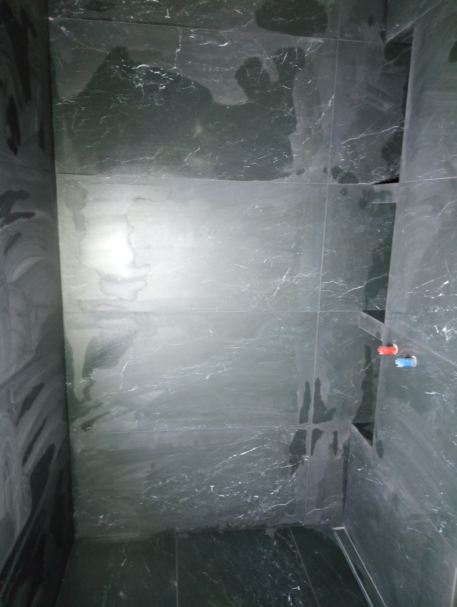 Wykończona kabina prysznicowa z ciemnymi, marmurkowymi płytkami, widoczne przyłącza wodne z oznaczeniem ciepłej i zimnej wody.