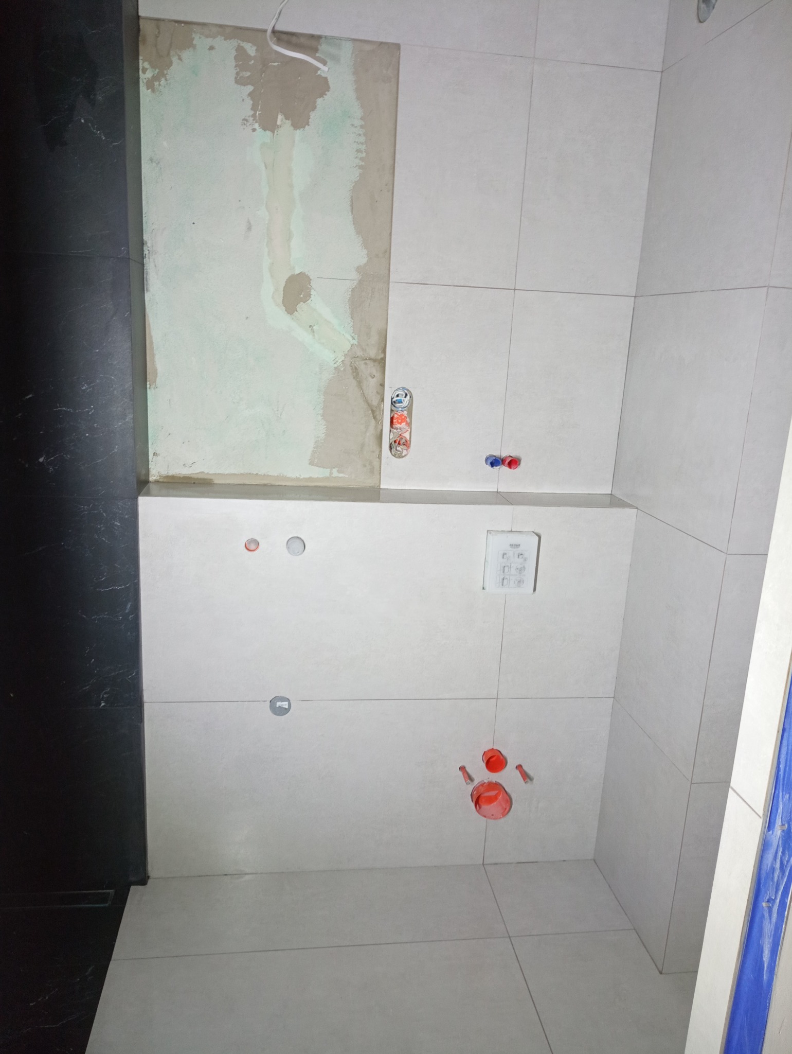 Ściana w trakcie układania jasnych, kwadratowych płytek ceramicznych. Widoczne otwory na instalacje, częściowo wykończona powierzchnia nad płytkami i czarna, marmurkowa podłoga.