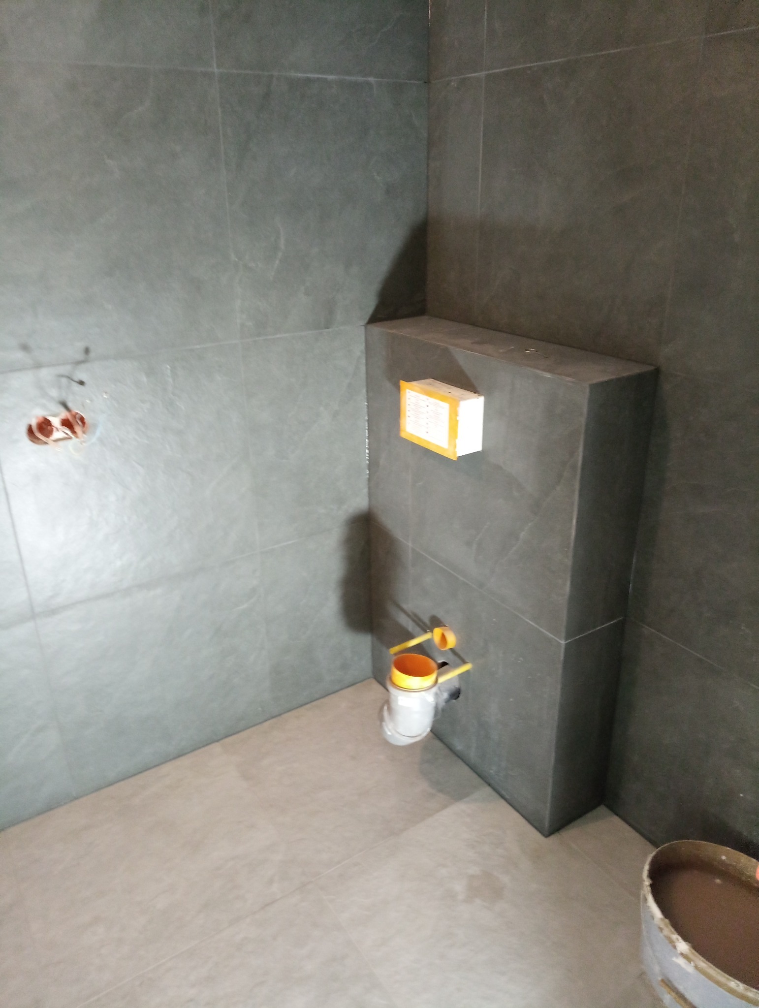 Szare płytki ceramiczne na ścianach i podłodze w trakcie remontu łazienki, widoczne elementy instalacji podtynkowej i wiadro z zaprawą.