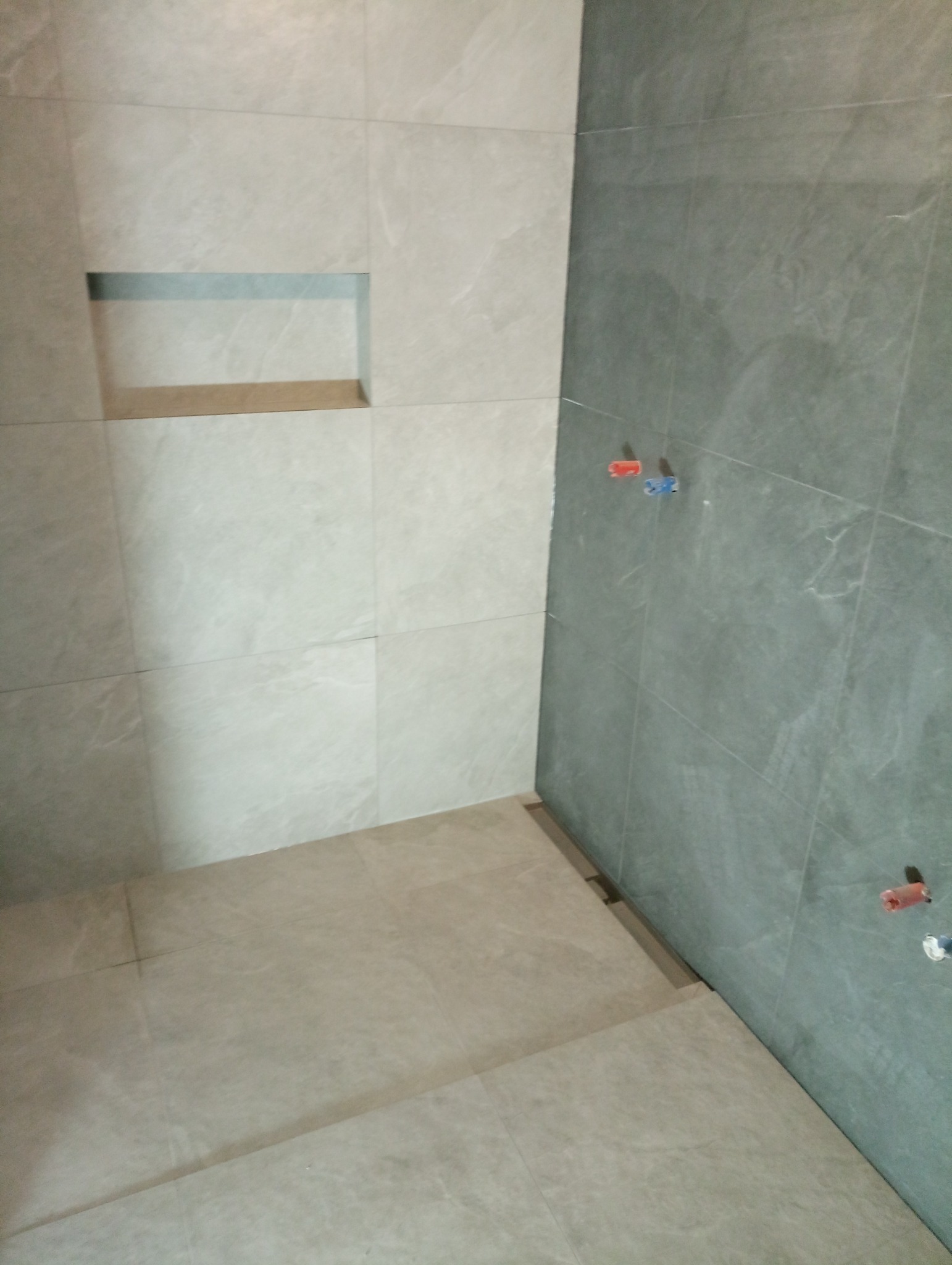 Wykończona łazienka z jasnymi płytkami na ścianach i podłodze, widoczna wnęka w ścianie i fragment ściany z szarymi płytkami z wyprowadzonymi rurami.