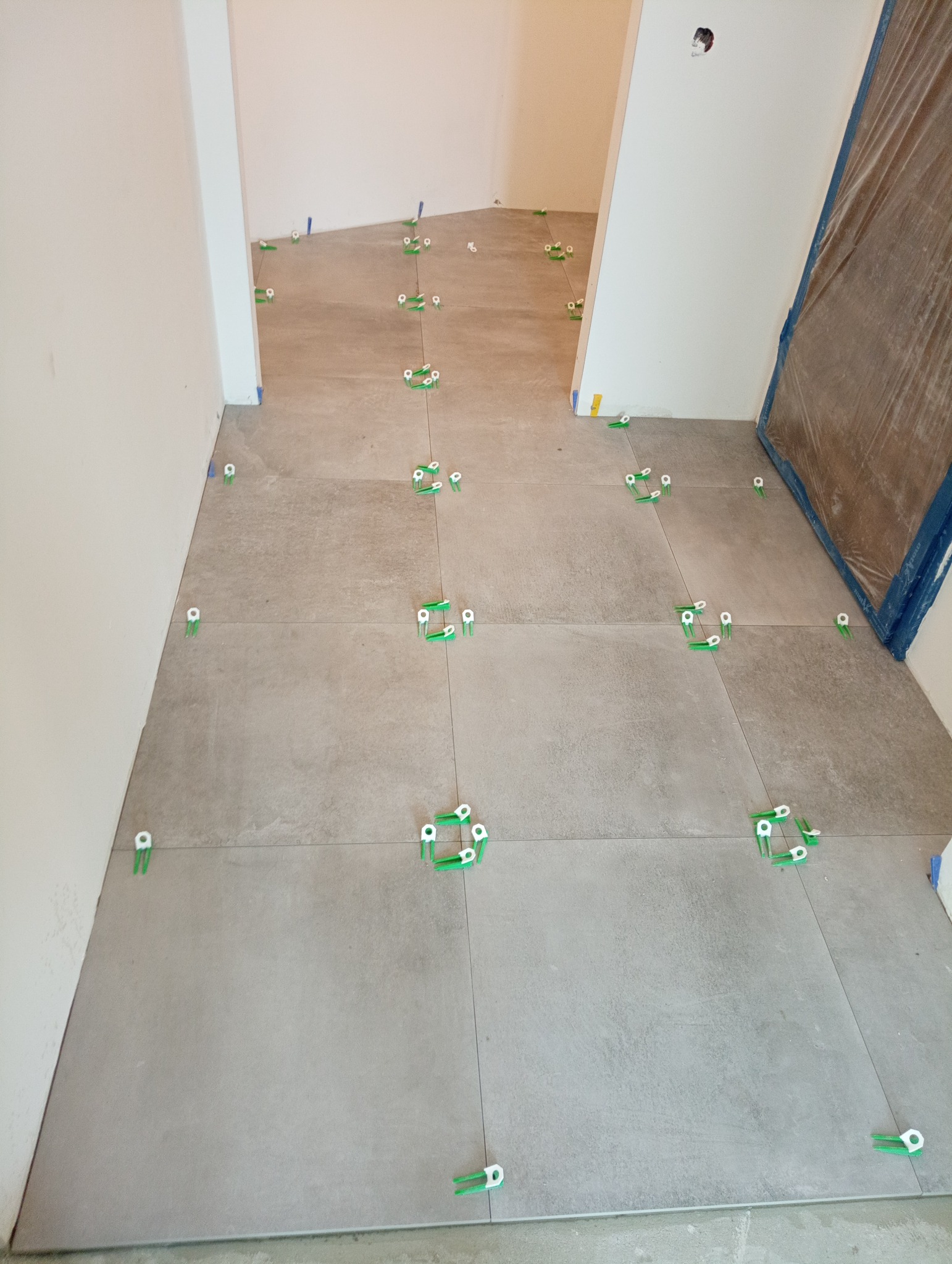 Ułożone płytki podłogowe w jasnoszarym odcieniu, z widocznymi zielonymi klipsami poziomującymi, w trakcie prac wykończeniowych w pomieszczeniu.