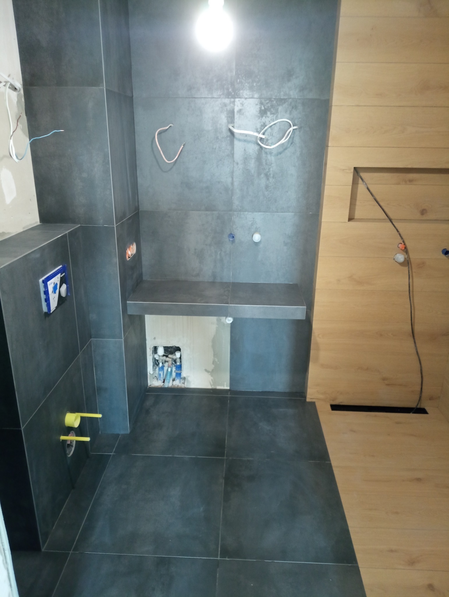 Wnętrze łazienki w trakcie remontu, widoczne szare płytki na ścianach i podłodze, drewniana boazeria, wystające kable elektryczne i rury hydrauliczne, brak armatury.