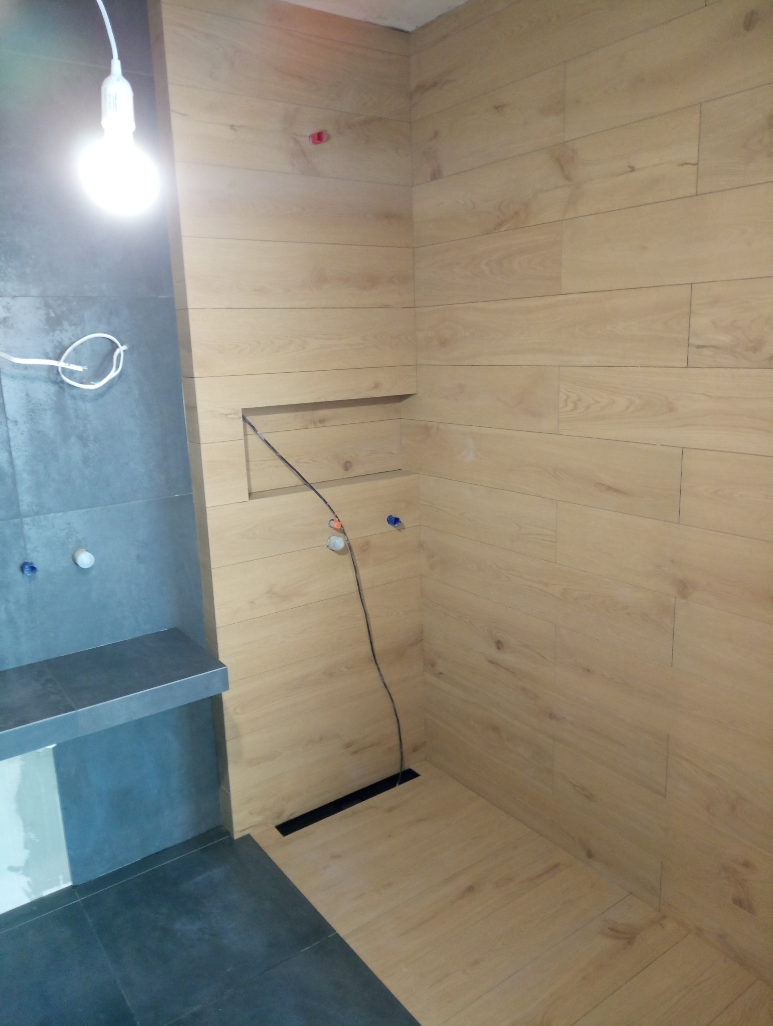 Płytki imitujące drewno na ścianach i podłodze w łazience w trakcie montażu, widoczna instalacja elektryczna i odpływ liniowy.
