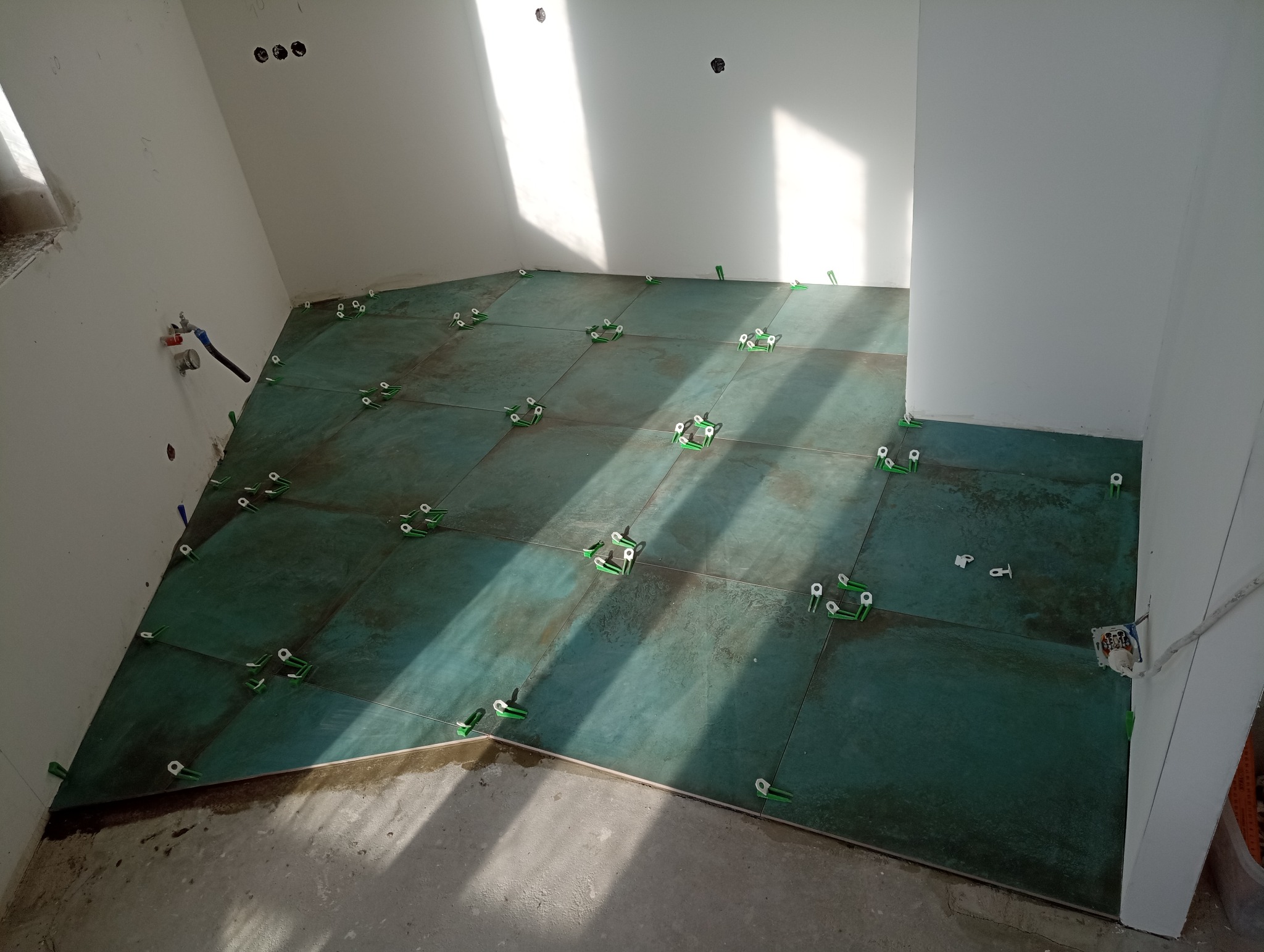 Układanie płytek podłogowych w łazience, widoczne zielone płytki z użyciem poziomujących klipsów i klinów, częściowo ukończone prace, surowy beton na podłodze przy ścianie.