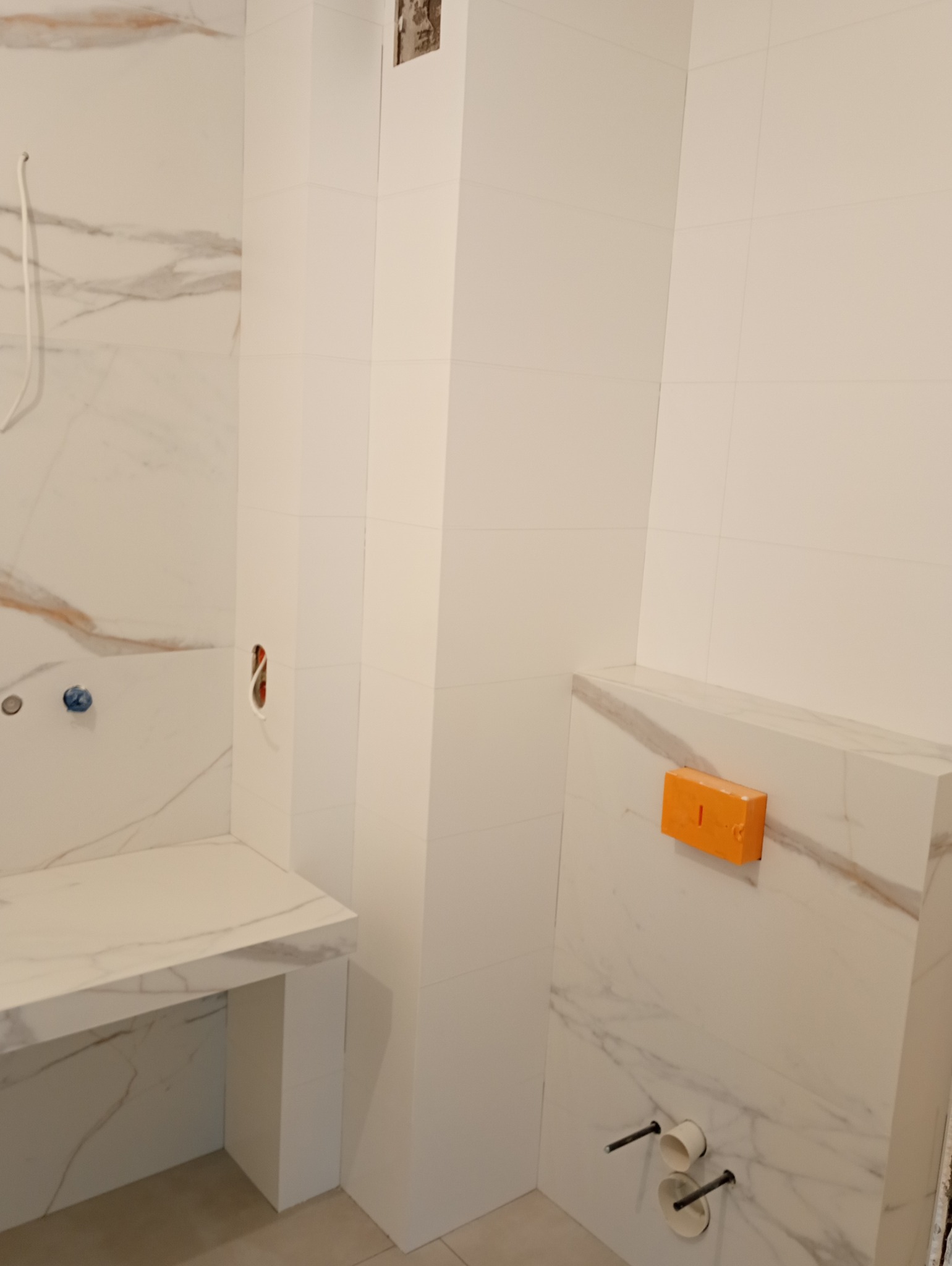 Białe płytki imitujące marmur na ścianach łazienki w trakcie montażu, widoczne wystające rury instalacji wodnej i elektrycznej, oraz poziomica przytwierdzona do ściany.