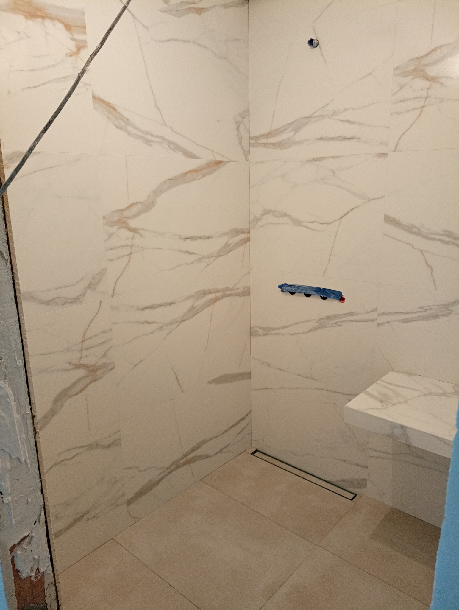 Wykończona łazienka z białymi płytkami imitującymi marmur, z zamontowanym odpływem liniowym i półką pod prysznicem, widoczne taśmy malarskie zabezpieczające armaturę.