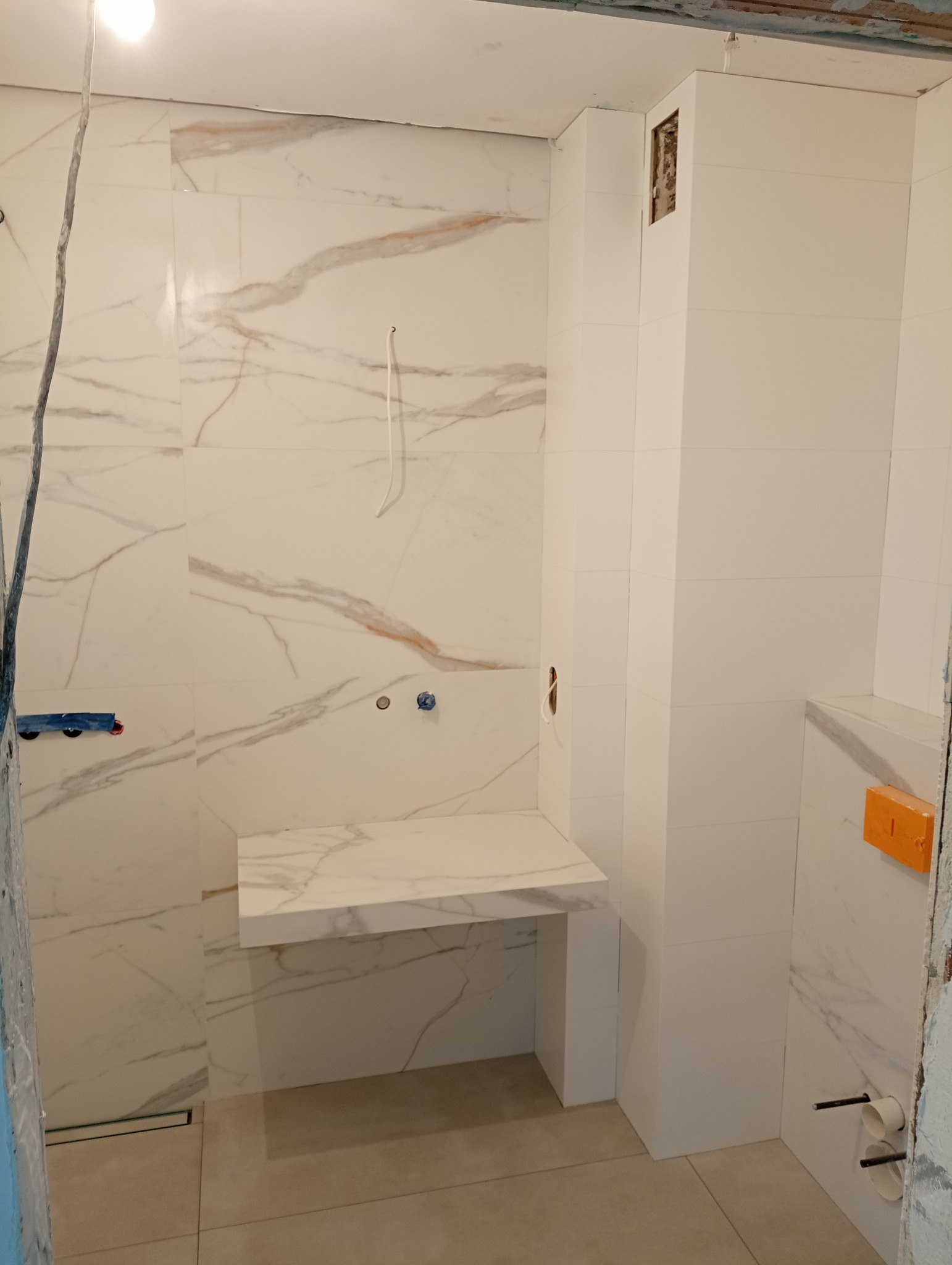 Łazienka w trakcie remontu: białe marmurowe płytki na ścianach, zabudowa pod umywalkę z tego samego materiału, widoczne instalacje wodne i elektryczne, surowy stan wykończenia.