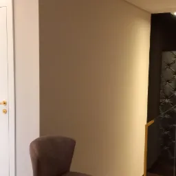 Krzesło tapicerowane w odcieniach szarości stoi na ciemnej, drewnianej podłodze przy białej ścianie i schodach z drewnianymi stopniami i złotą balustradą.