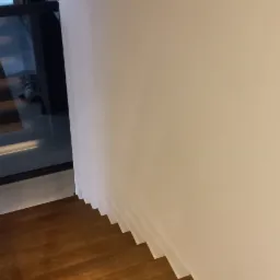 Widok z góry na drewniane schody z panelami, z widocznym fragmentem okna i białej ściany, brak poręczy.