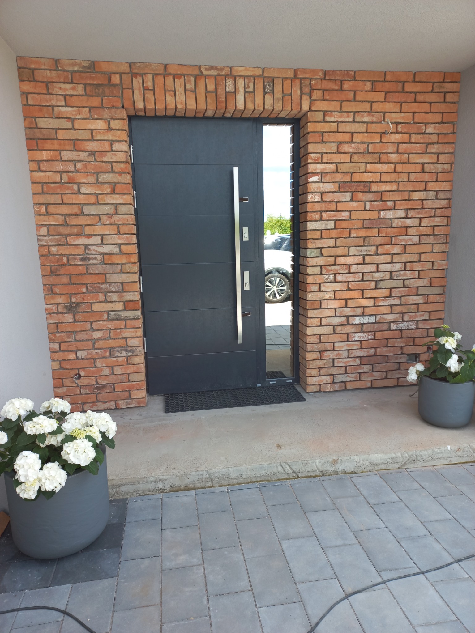 Ciemnoszare drzwi wejściowe z długim, srebrnym uchwytem, otoczone czerwoną cegłą, z donicami z białymi kwiatami po obu stronach, na zewnątrz budynku.