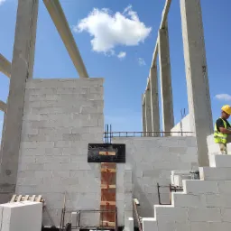 Wznoszenie ściany z białych bloczków na budowie domu, widoczne betonowe słupy konstrukcyjne, pracownik w kasku i kamizelce mierzy poziom.